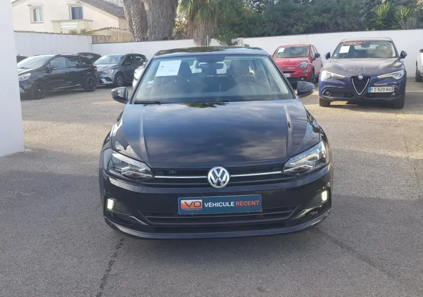 Vue avant d'une Volkswagen Polo noire 2018 avec feux diurnes allumés sur un parking extérieur.