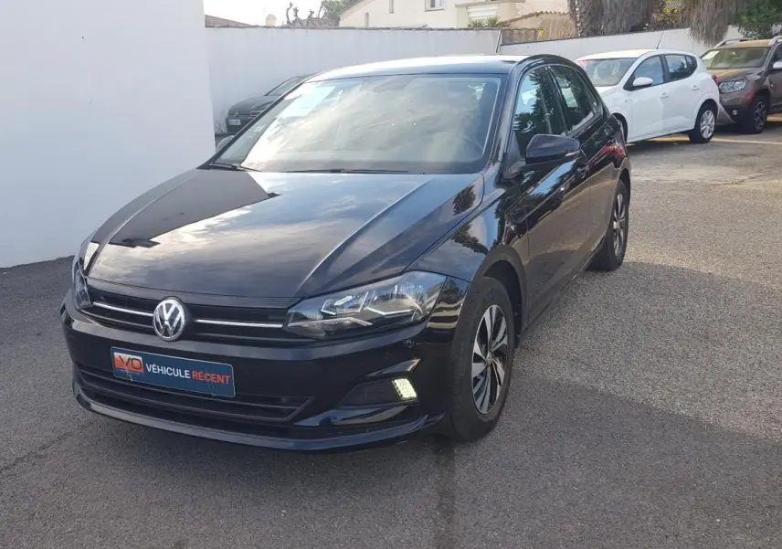 Volkswagen Polo noire vue 3/4 avant droit avec jantes alliage et feux diurnes allumés sur parking.