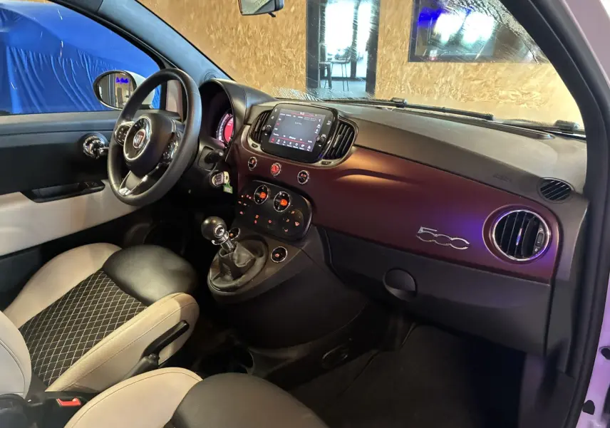 Vue intérieure côté conducteur de la Fiat 500 blanche, avec tableau de bord violet et volant multifonction.