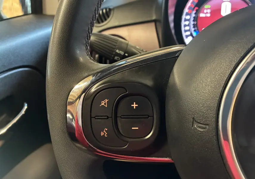 Gros plan sur les commandes de volume au volant noir de la Fiat 500 blanche, avec tableau de bord partiellement visible.