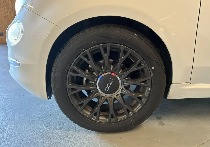 Gros plan sur la roue avant gauche d'une FIAT 500 blanche de 2021, mettant en valeur la jante noire au design fin et le logo central.