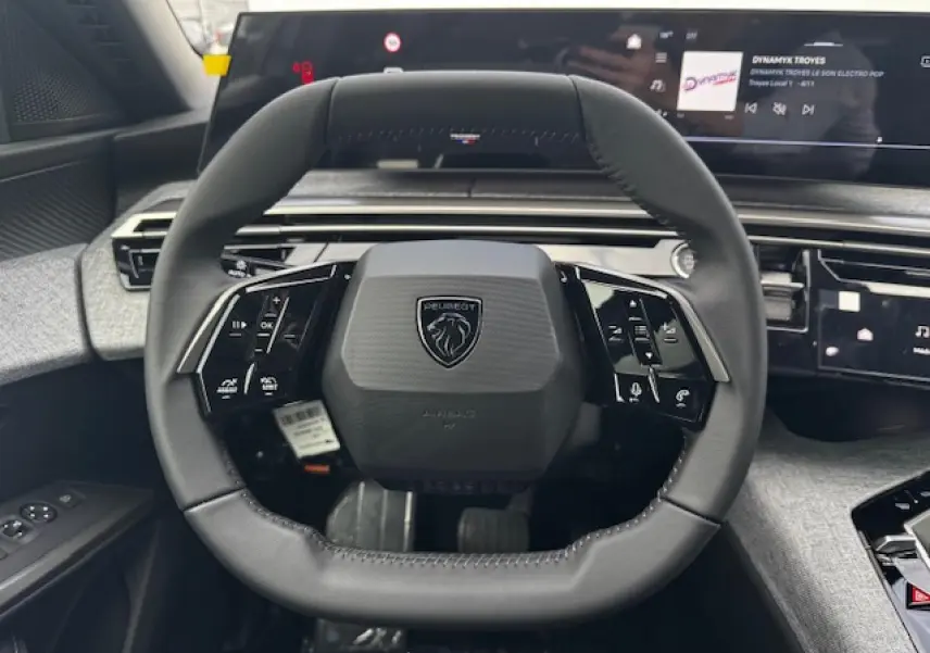 Volant compact cuir noir avec logo Peugeot, tableau de bord numérique panoramique et habillage tissu gris clair visible depuis le poste de conduite