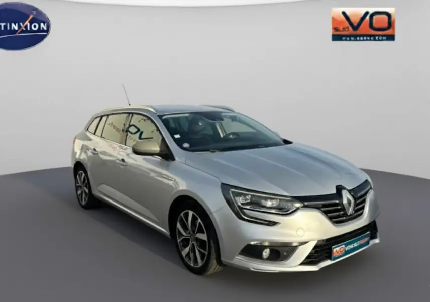 Renault Mégane Break gris clair vue 3/4 avant droit, avec jantes alliage et barres de toit visibles.