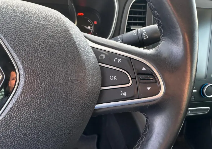 Gros plan sur le volant cuir noir de la Renault Mégane Break gris clair, avec commandes intégrées et tableau de bord partiel visible.