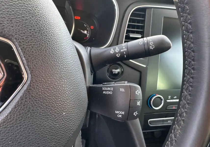 Gros plan sur le volant cuir et commandes audio d'une Renault Mégane Break gris clair, tableau de bord et écran tactile visibles.
