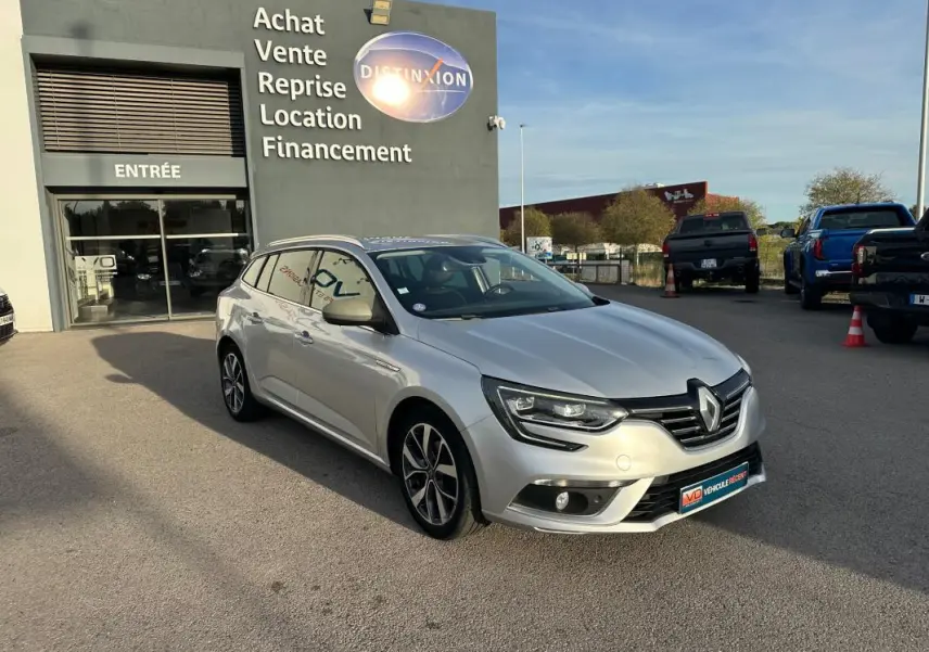 Renault Mégane Break gris clair vue 3/4 avant droit, avec jantes alliage 18 pouces et barres de toit visibles.