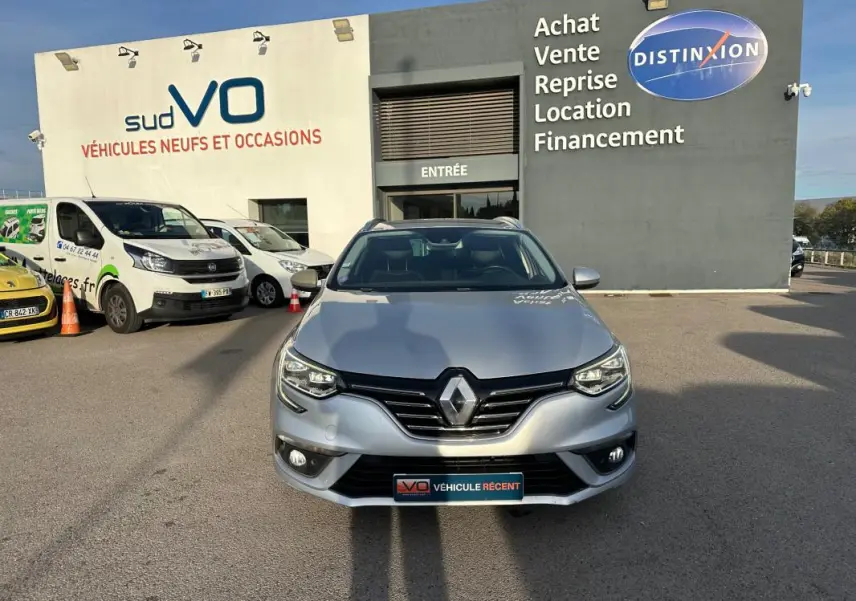 Renault Mégane Break gris clair vue de face, avec barres de toit et phares allumés sur parking de concession.