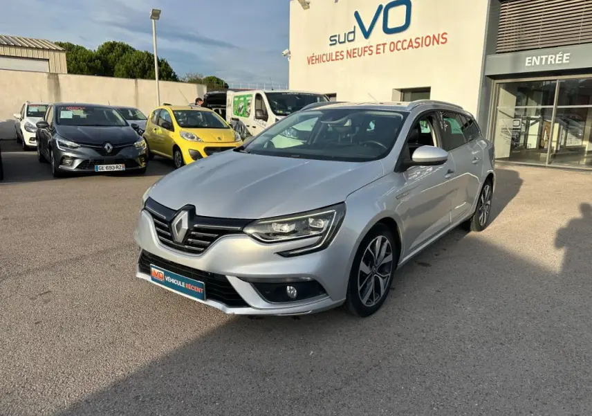 Renault Mégane Break gris clair vue 3/4 avant droit avec jantes alliage 18 pouces, barres de toit et toit ouvrant électrique.