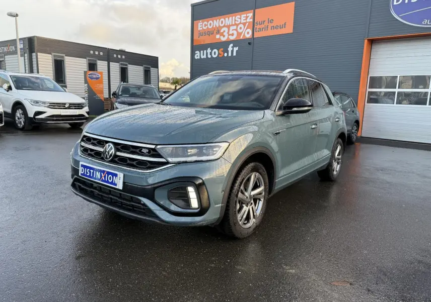 Volkswagen T-Roc bleu pétrole métallisé en 3/4 avant droit sur parking devant concession avec panneau promotionnel.