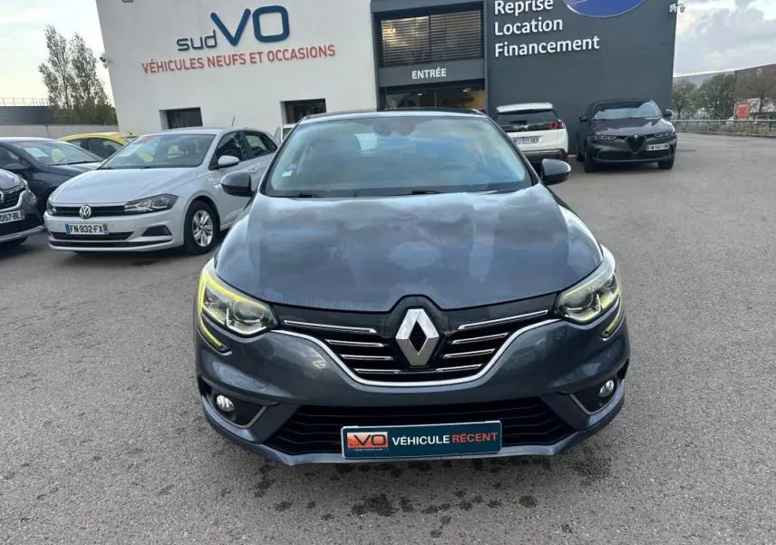 Vue frontale d'une Renault Mégane IV gris foncé avec phares allumés, garée devant un concessionnaire sudVO.