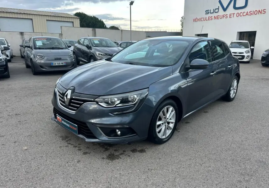 Renault Mégane IV gris foncé vue 3/4 avant droit, avec jantes alliage et phares LED allumés, sur parking extérieur.