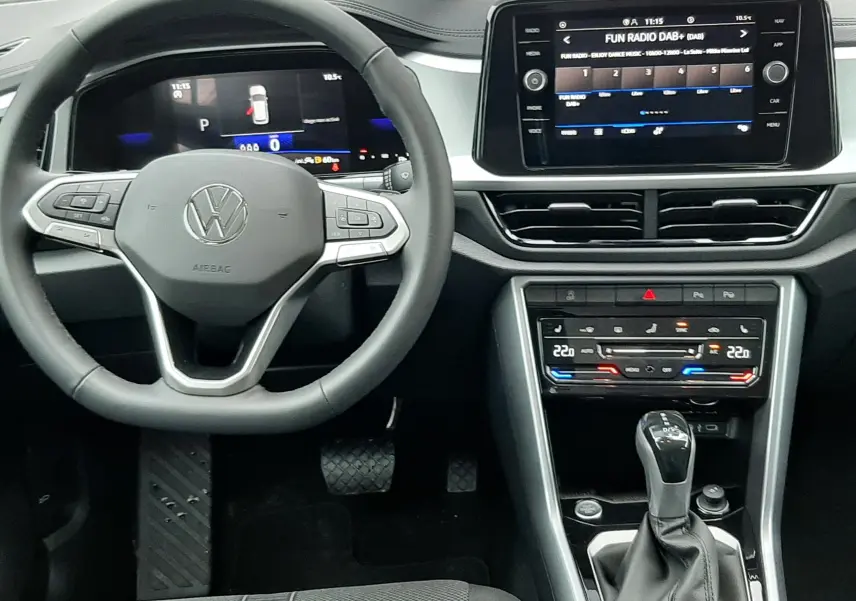 Vue intérieure centrée sur le volant et la console centrale du Volkswagen T-Roc 1.5 TSI gris, avec écran tactile et levier de vitesse automatique.