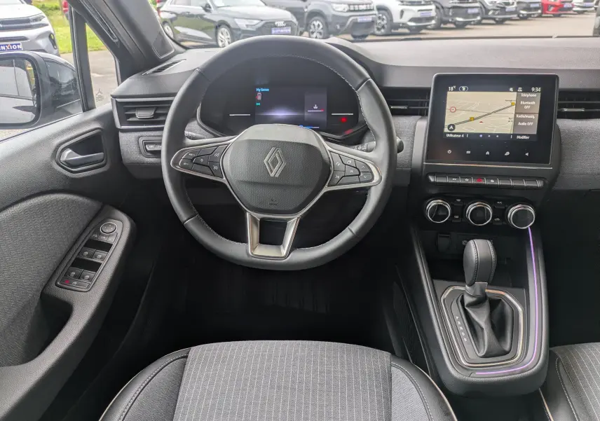 Vue intérieure avant centrée sur le volant et tableau de bord de la Renault Clio hybride 2025 en noir étoile métal, avec écran tactile et levier de vitesses.