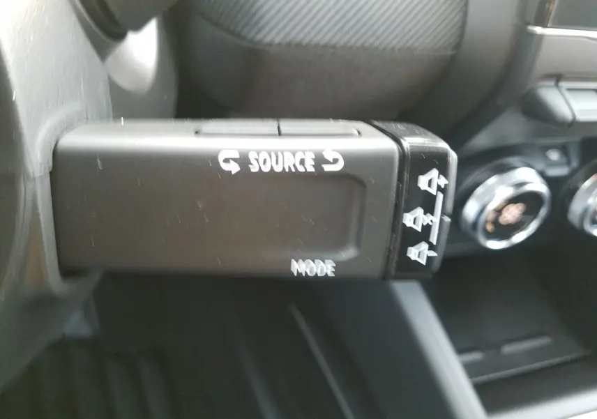 Gros plan sur la commande au volant de la Renault Clio V hybride 2025, boutons source et mode visibles en noir.