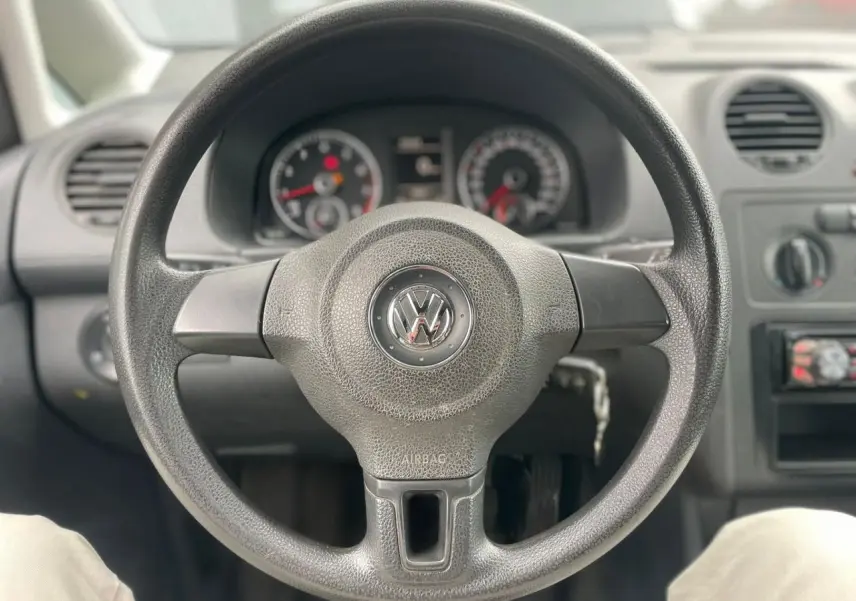 Vue rapprochée du volant noir de la Volkswagen Caddy 2014 avec tableau de bord et commandes visibles en arrière-plan.