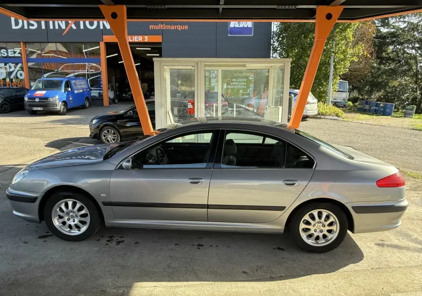 Profil côté gauche d'une Peugeot 607 grise stationnée sous un abri orange avec des jantes alliage visibles.