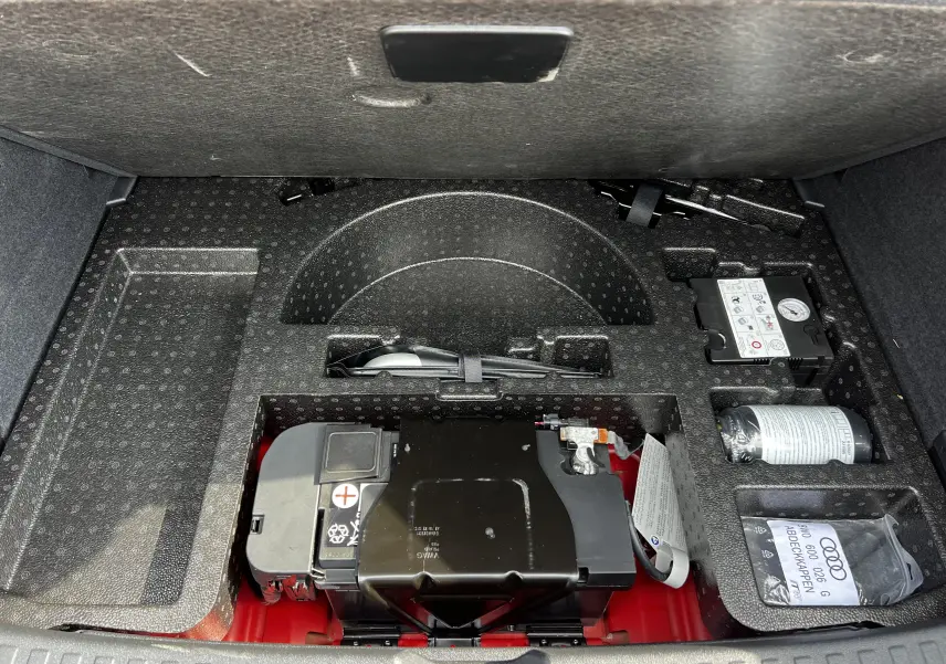 Compartiment de rangement sous le coffre d'une Audi RS3 Sportback rouge, montrant batterie et kit de réparation pneus.