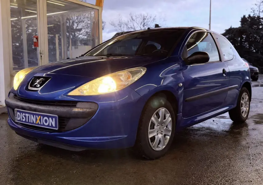 Peugeot 206+ bleu vue 3/4 avant droit avec phares allumés, garée devant un bâtiment vitré.