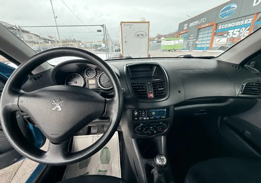 Vue intérieure avant de la Peugeot 206+ bleue, montrant le volant, tableau de bord et levier de vitesse manuel.