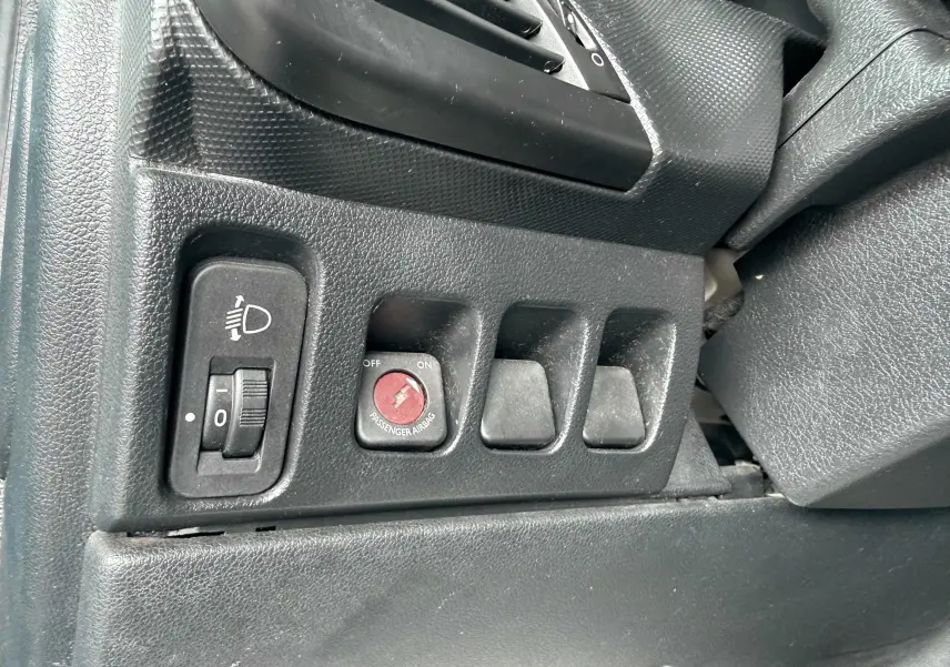 Gros plan sur les commandes noires côté gauche du tableau de bord d'une Peugeot 206+ bleue, avec réglage des phares et airbag passager.