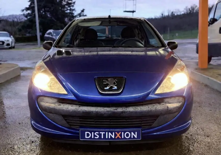Vue avant d’une Peugeot 206+ bleue de 2011 avec phares allumés et plaque Distinxion visible.