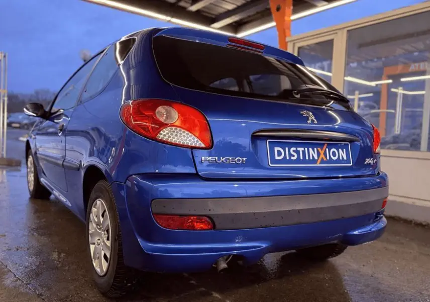 Vue 3/4 arrière gauche d'une Peugeot 206+ bleue garée sous un abri avec plaque Distinxion visible.