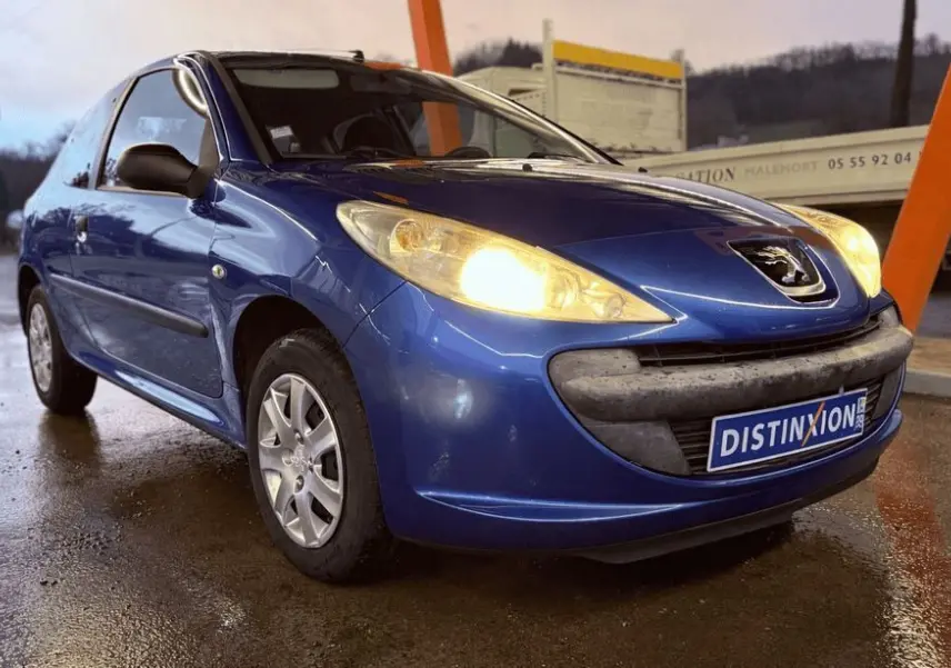 Peugeot 206+ bleu vue 3/4 avant droit avec phares allumés sur sol mouillé, devant structure orange.