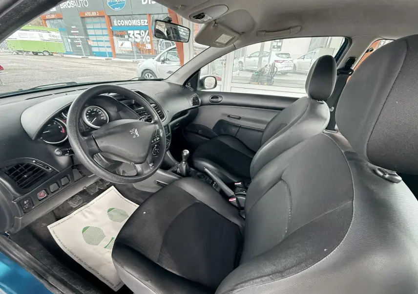 Vue intérieure côté conducteur d'une Peugeot 206+ bleue de 2011 avec sièges tissu noir et volant cuir.