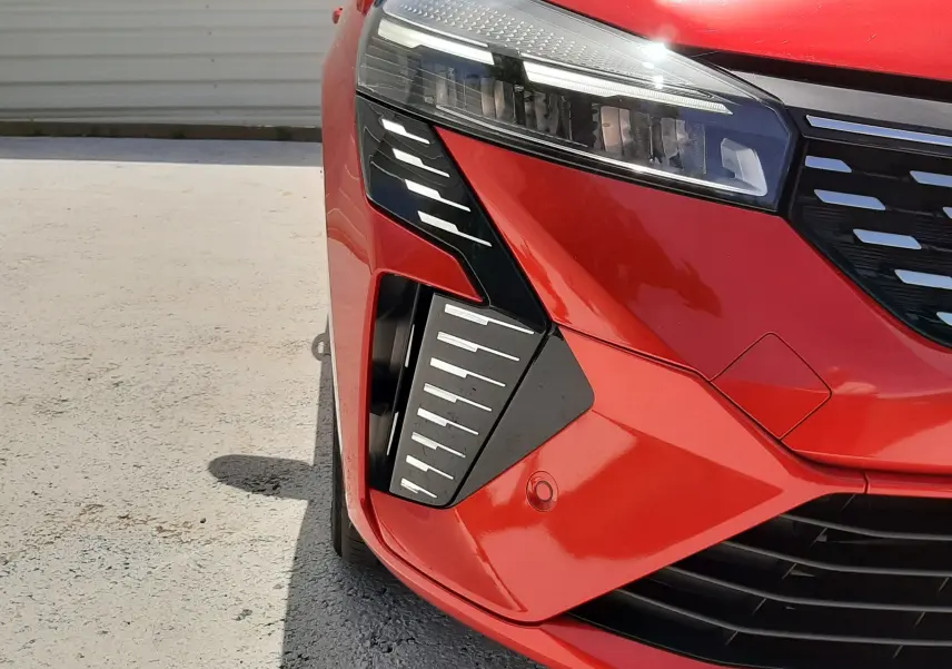 Gros plan sur l'avant droit de la Renault Clio V rouge flamme métal, mettant en valeur son phare LED et la calandre noire moderne.