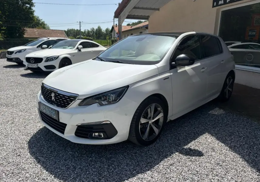 Peugeot 308 GT-Line blanche vue en 3/4 avant droit, avec jantes alliage et toit noir contrasté, stationnée sur gravier.