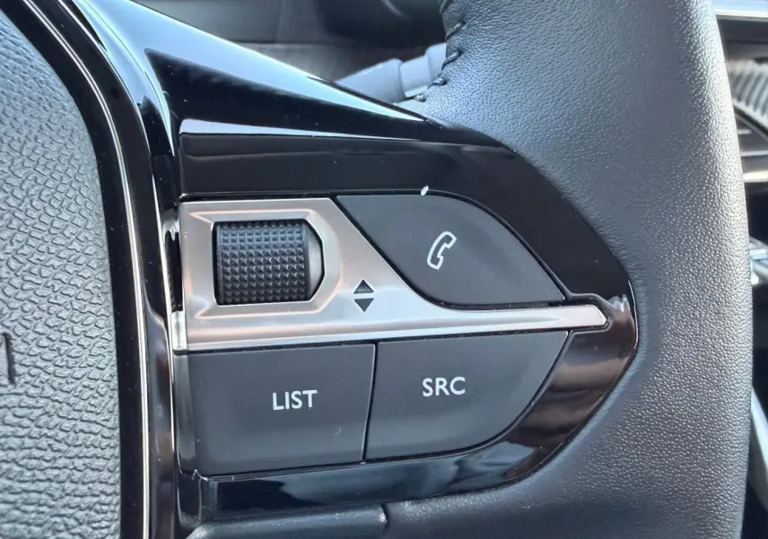 Gros plan sur les commandes audio au volant noir de la Peugeot 208 1.2 100ch Gris Artense, montrant molette et boutons SRC, LIST.