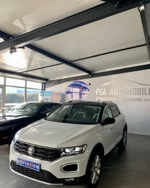 Volkswagen T-Roc blanc en 3/4 avant droit dans un showroom avec calandre noire et phares LED allumés.
