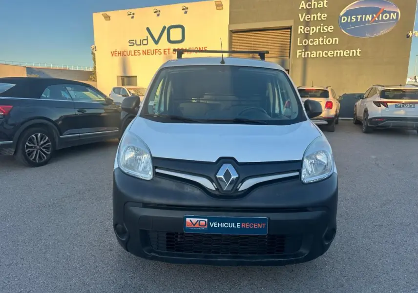 Vue frontale d'un Renault Kangoo Express blanc avec pare-chocs noir mat, stationné devant un bâtiment commercial.