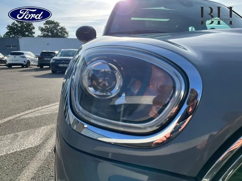 Gros plan sur le phare avant gauche chromé d'une MINI Countryman Moonwalk Grey, avec reflet du ciel et environnement urbain.