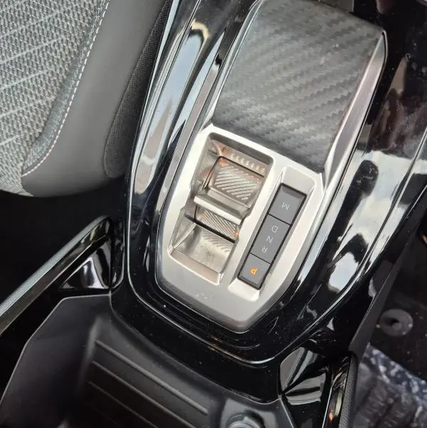 Gros plan sur la console centrale noire brillante de la Peugeot 208 Hybrid 110 e-DCS6 Allure 2025 avec levier de vitesses automatique et prise 12V.