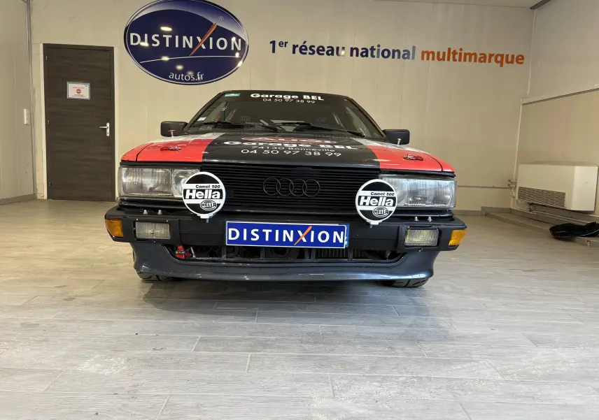 Vue frontale d'une Audi Coupé GT noire avec capot rouge, équipée de deux phares additionnels Hella et stickers de rallye.