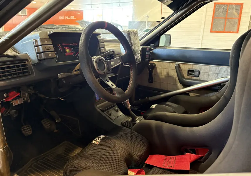 Vue intérieure côté conducteur de l'Audi Coupé GT noir de 1983, avec sièges baquets, harnais rouge et volant course.