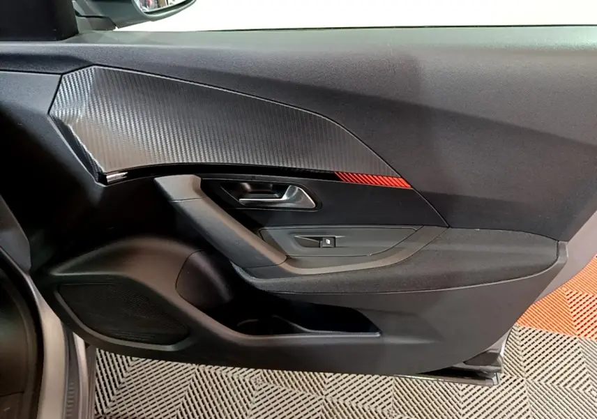 Vue intérieure du panneau de porte avant gauche du Peugeot 2008 gris clair, avec insert imitation carbone et bande rouge décorative.