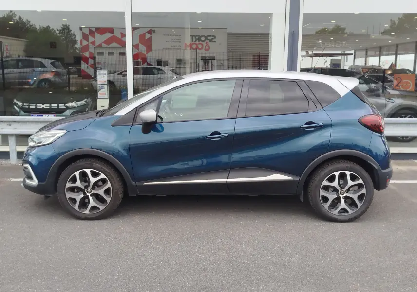 Profil droit d’un Renault Captur 2018 bleu marine fumé avec toit gris platine et jantes bi-ton, garé devant une vitrine.