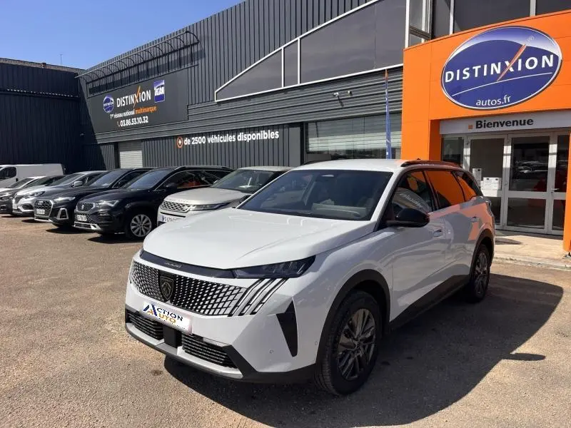 Peugeot 5008 Hybrid blanc Okénite en 3/4 avant droit, avec calandre distinctive et jantes alu, devant un concessionnaire.