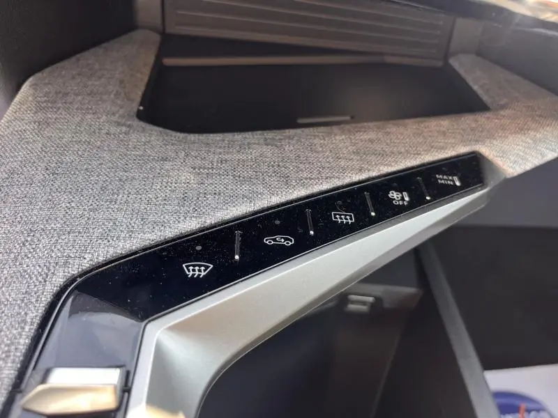 Gros plan sur la console centrale avec commandes tactiles noires et tissu gris clair dans un Peugeot 5008 Hybrid blanc.