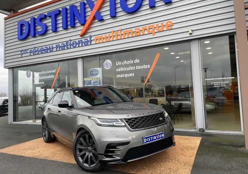 Range Rover Velar D300 gris Silicon Silver en 3/4 avant droit devant un showroom Distinxion, avec jantes foncées et phares LED allumés.