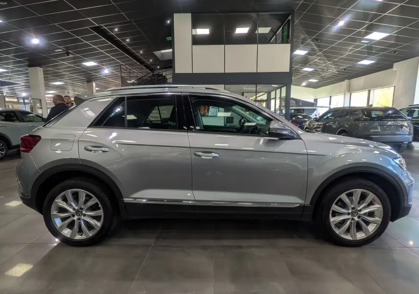 Profil côté droit d'un Volkswagen T-Roc gris pyrite 2024 en showroom, avec jantes alliage 18 pouces et toit noir.