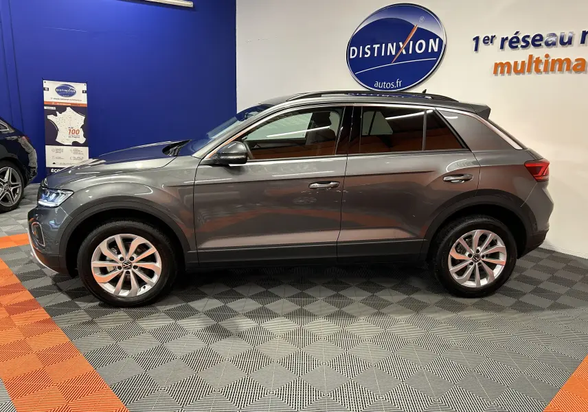 Profil droit d'un Volkswagen T-Roc gris Indium Metal 2025 en showroom, avec jantes alliage et vitres teintées.