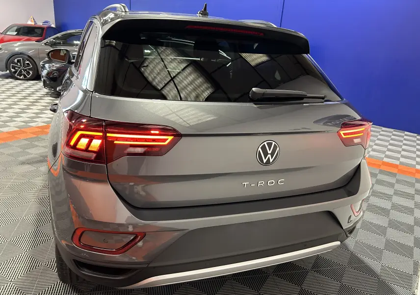 Vue 3/4 arrière d’un Volkswagen T-Roc gris Indium Metal avec feux arrière allumés dans un showroom.