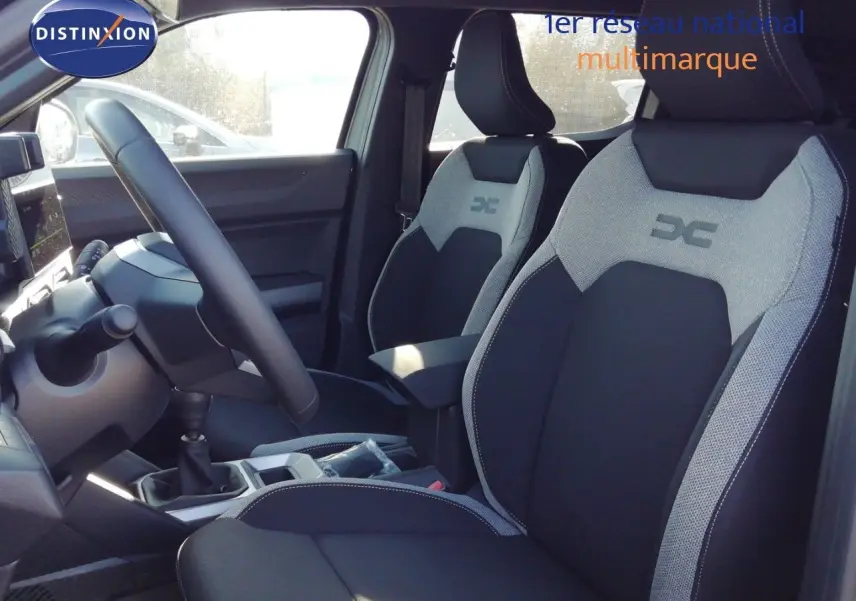 Vue intérieure côté conducteur du Dacia Duster 2026, sièges tissu gris et noir avec logo DC brodé, volant multifonction.