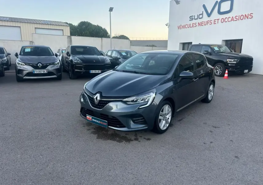 Renault Clio gris foncé vue 3/4 avant droit sur parking devant concession Sud VO, avec phares allumés et jantes alliage.