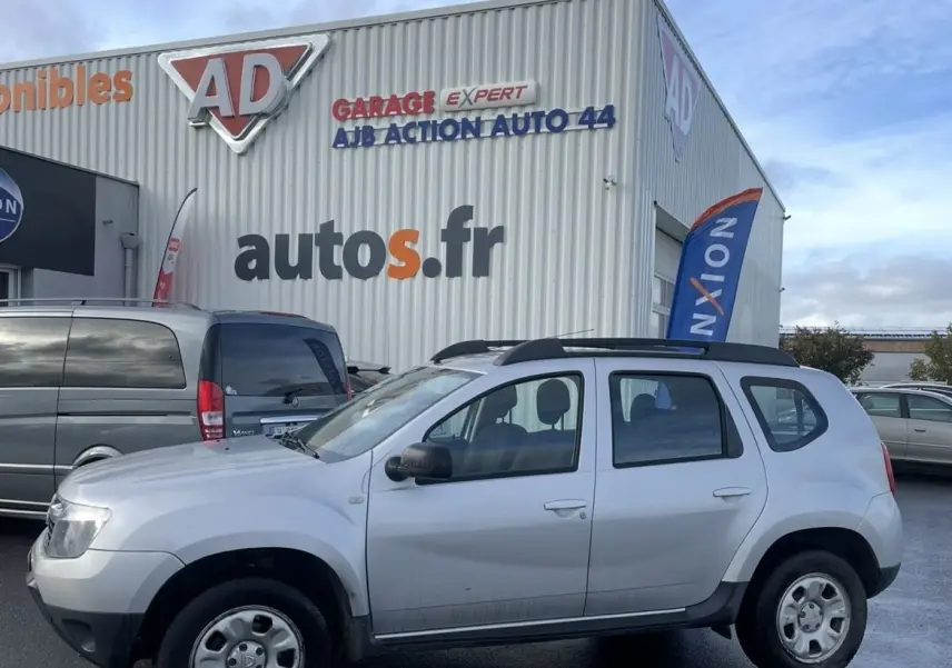 Vue de profil côté gauche d'un Dacia Duster gris avec barres de toit et enjoliveurs, stationné devant un garage.