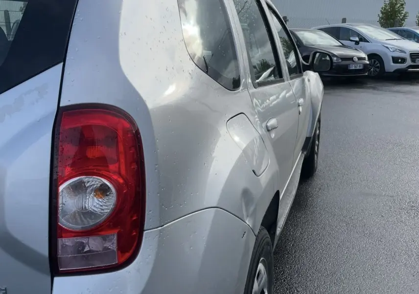 Vue 3/4 arrière gauche d'un Dacia Duster gris avec barres de toit et attelage sous un ciel nuageux.