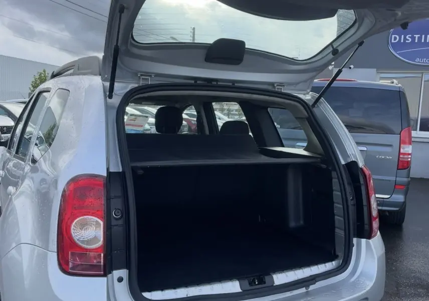 Vue arrière ouverte d'un Dacia Duster gris 2011 avec attelage et coffre spacieux visible sous un ciel nuageux.
